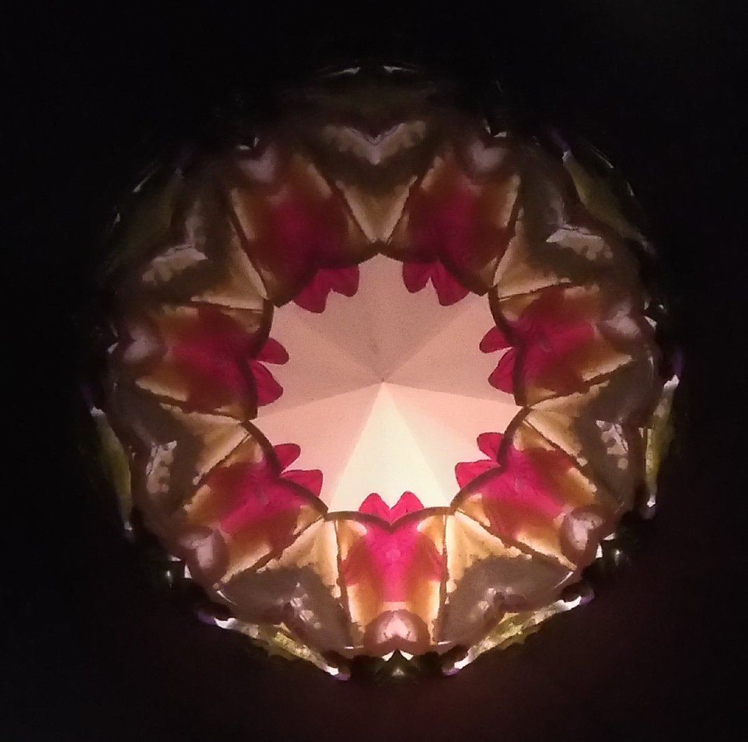 Kaléidoscopie florale. 7 pétales roses sont reliés par une couronne vert sombre. Le coeur est un rond lumineux avec une forme de prisme qui pointe vers la personne qui regarde. Les pétales se terminent en forme de cœur qui pointe vers l’intérieur du cercle.