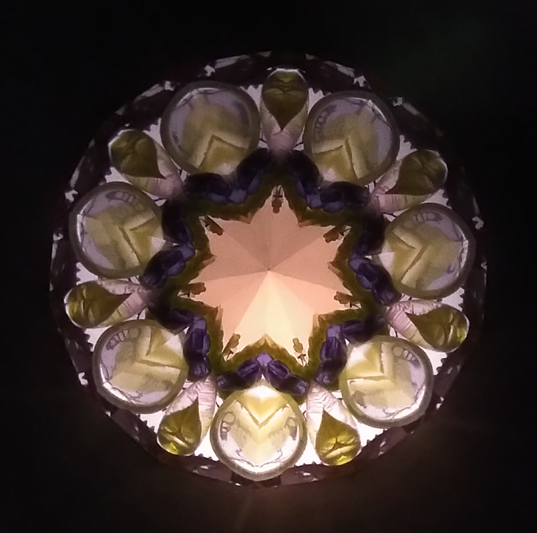 Kaléidoscopie, cœur lumineux en forme d’étoile à 7 branches, doublé d’une étoile plus sombre. Sur l'extérieur, une fleur à 7 pétales vert-dorés avec des gouttes entre chaque pétale.