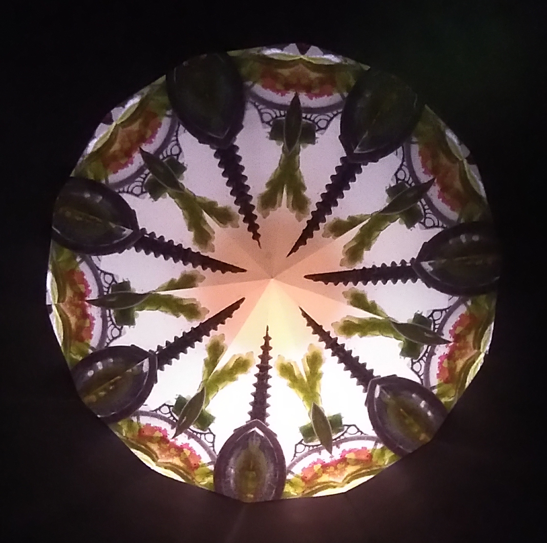 Kaléidoscopie, cœur lumineux en forme d’étoile à 7 branches. Les bords des branches sont vert bouteille. Chaque branche se termine par une forme qui évoque une fleur de lys vert bouteille. Sur le contour, 7 demis-cercles décorés d’un motif vert et marron évoquant une salade feuilel de chêne. Entre chaque demi-cercle, des formes ovoïdes se terminant par une corne en spirale pointent vers le centre de la kaléidoscopie.