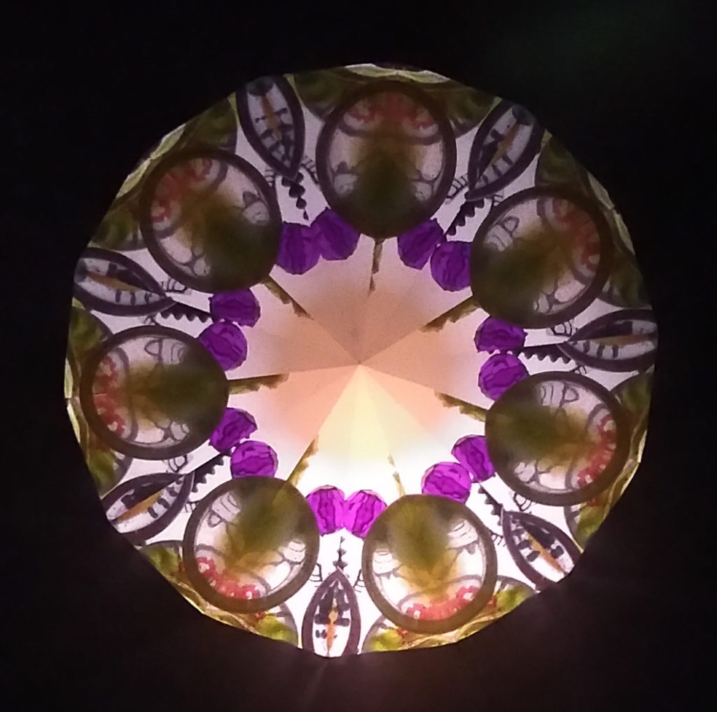 Kaléidoscopie, cœur lumineux de forme arrondie. 7 traits vert foncé ornent le cœur. Celui-ci est entouré d’une couronne de 14 perles violettes. Une frise de cercles décoré de vert et de orange se trouve sur le pourtour du premier cercle. Enfin, des demi-cercles tournés vers l’extérieur terminent le motif.