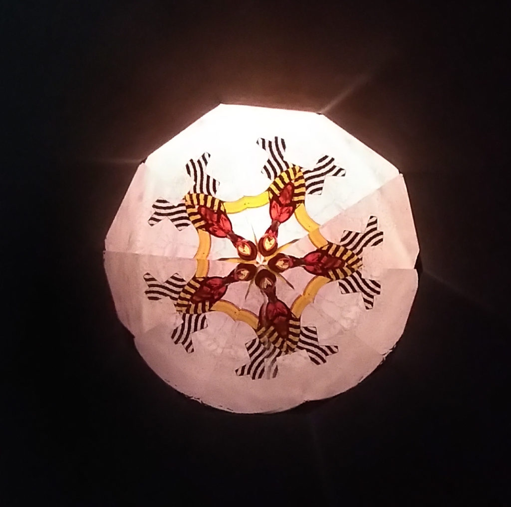 Kaléidoscopie. Au centre, une étoile à 5 branches orange et jaune. Un pentagone jaune l’encadre. Les branches se terminent par des fleurs en forme de clochette rouge. Celles-ci sont prolongées par un V rayé noir et blanc. Le reste est lumineux.