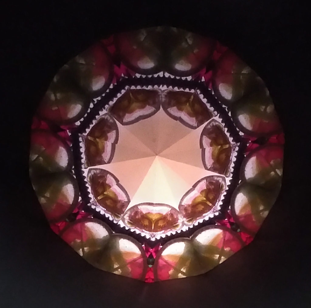 Kaléidoscopie octogonale. Le centre est lumineux. Le tour noir est fin et doublé d’une couronne brune-ocre entouré de perles blanches lumineuses, 2 autres rangs de perles, un noir et un blanc. Sur le pourtour, un décor de feuilles vertes et- de papillons roses.