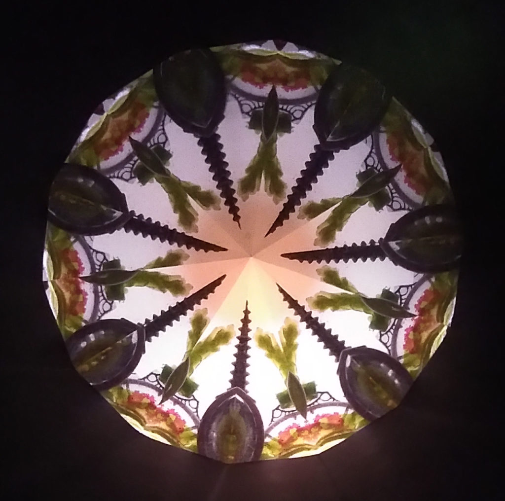 Kaléidoscopie, cœur lumineux en forme d’étoile à 7 branches. Les bords des branches sont vert bouteille. Chaque branche se termine par une forme qui évoque une fleur de lys vert bouteille. Sur le contour, 7 demis-cercles décorés d’un motif vert et marron évoquant une salade feuille de chêne. Entre chaque demi-cercle, des formes ovoïdes se terminant par une corne en spirale pointent vers le centre de la kaléidoscopie.